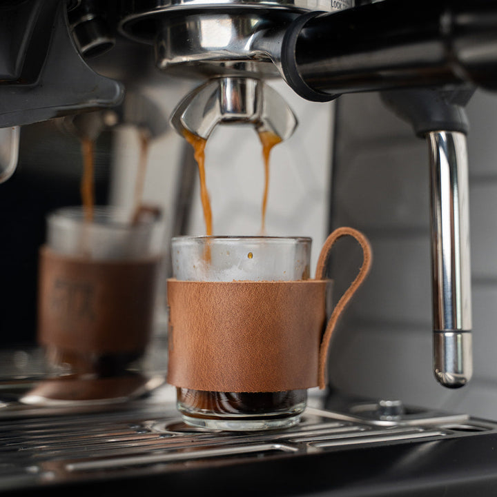 Espresso being brewed into a glass cup with a leather sleeve with a stamped debossed Texas A&amp;M Aggies on an espresso machine.