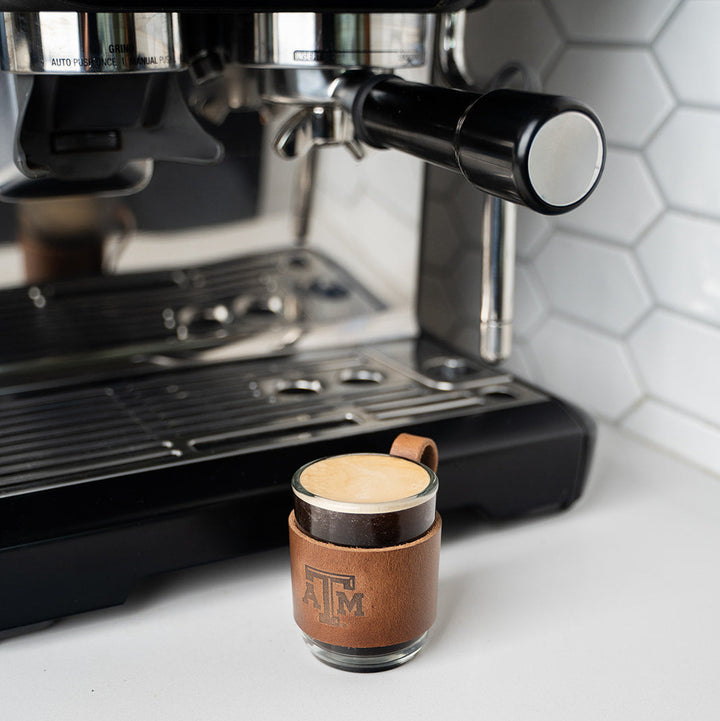 Espresso machine with a small leather cup with a leather stamped debossed Texas A&amp;M Aggies logo on a white countertop.