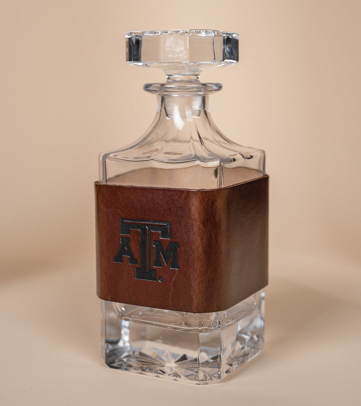 Glass decanter with leather sleeve featuring a Texas A&amp;M stamped debossed logo on a beige background