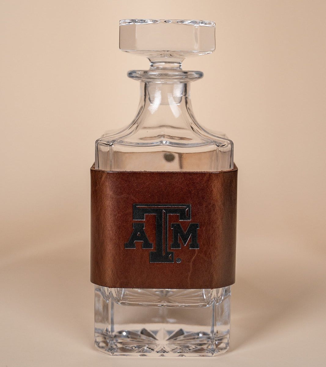 Glass decanter with leather wrap with a Texas A&amp;M stamped debossed logo on a beige background