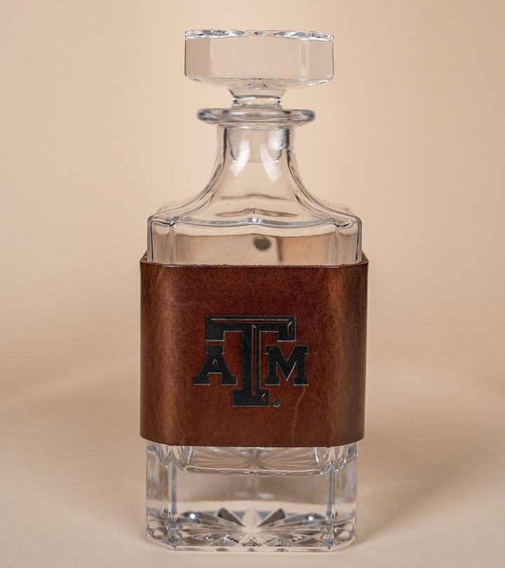 Glass decanter with leather wrap with a Texas A&amp;M stamped debossed logo on a beige background