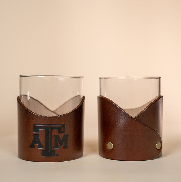 Two glass tumblers with brown leather sleeves featuring a stamped debossed Texas A&amp;M Aggies logo on a beige background