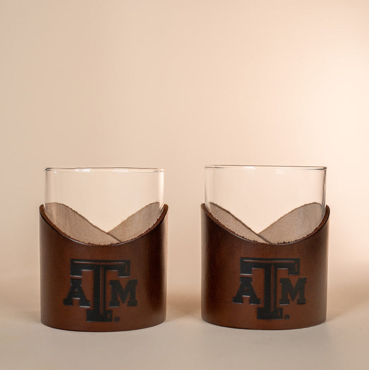 Two glass tumblers with brown leather sleeves featuring a stamped debossed Texas A&amp;M Aggies logo on a beige background