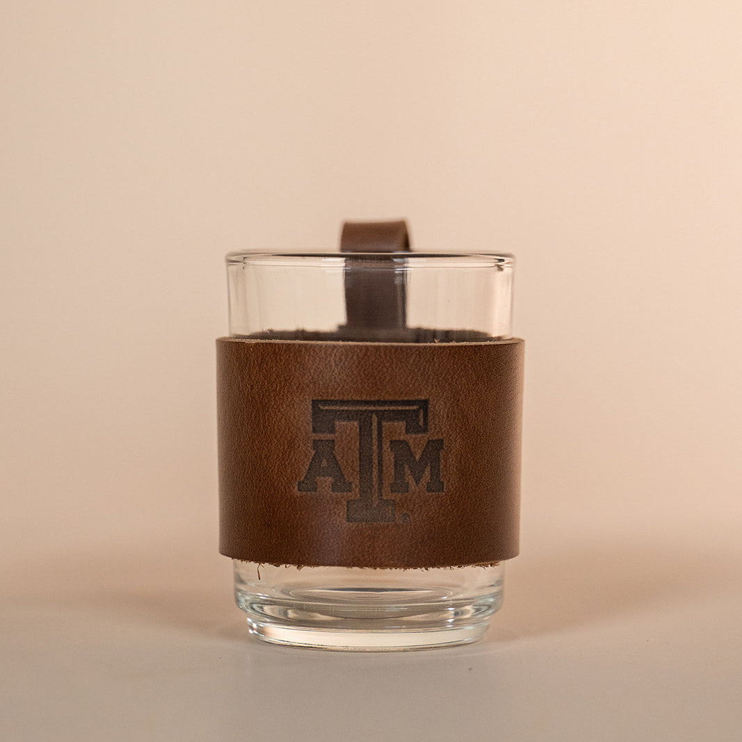Clear glass with a brown leather sleeve featuring a Texas A&amp;M stamped debossed logo on a beige background