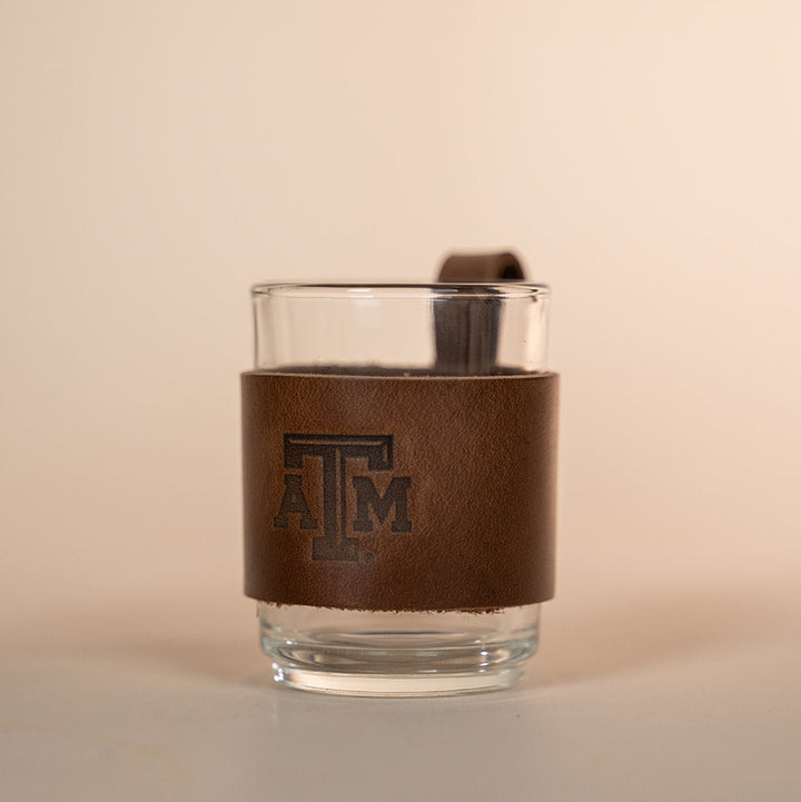 Clear glass with a brown leather sleeve featuring a Texas A&amp;M stamped debossed logo on a beige background