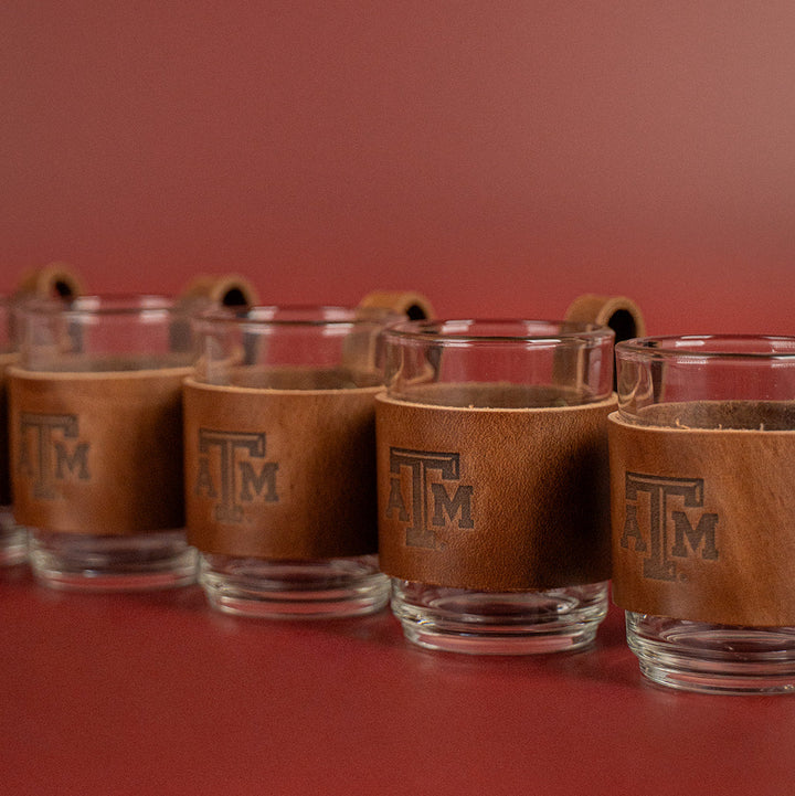 6 clear glasses with a brown leather sleeve featuring a Texas A&amp;M stamped debossed logo on a beige background