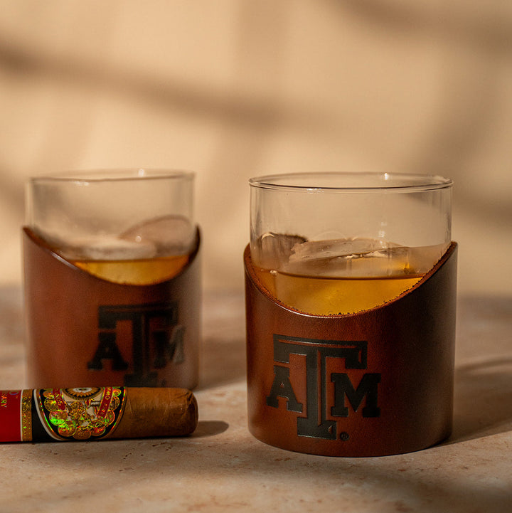 Two glass tumblers with brown leather sleeves featuring a stamped debossed Texas A&amp;M Aggies logo on a beige background with bourbon and a cigar
