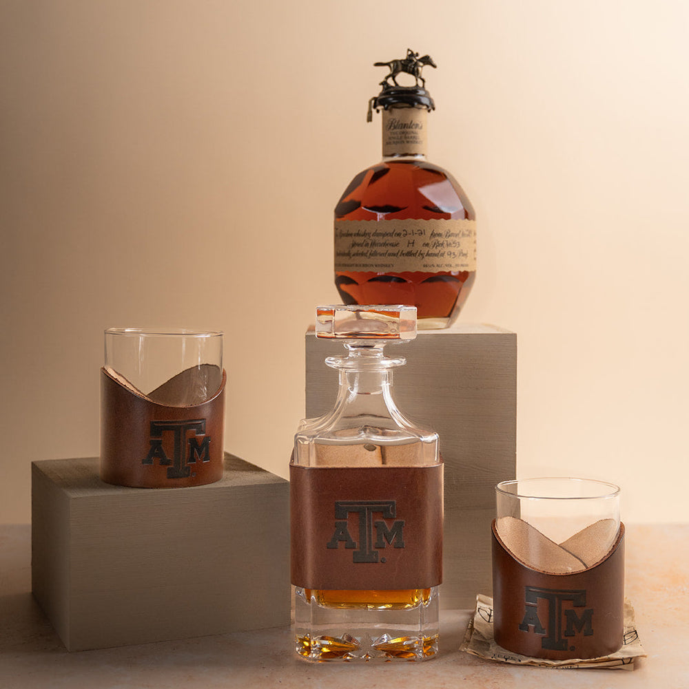 Two glass tumblers with brown leather sleeves featuring a stamped debossed Texas A&M Aggies logo on a beige background with Blanton's bourbon and a leather decanter