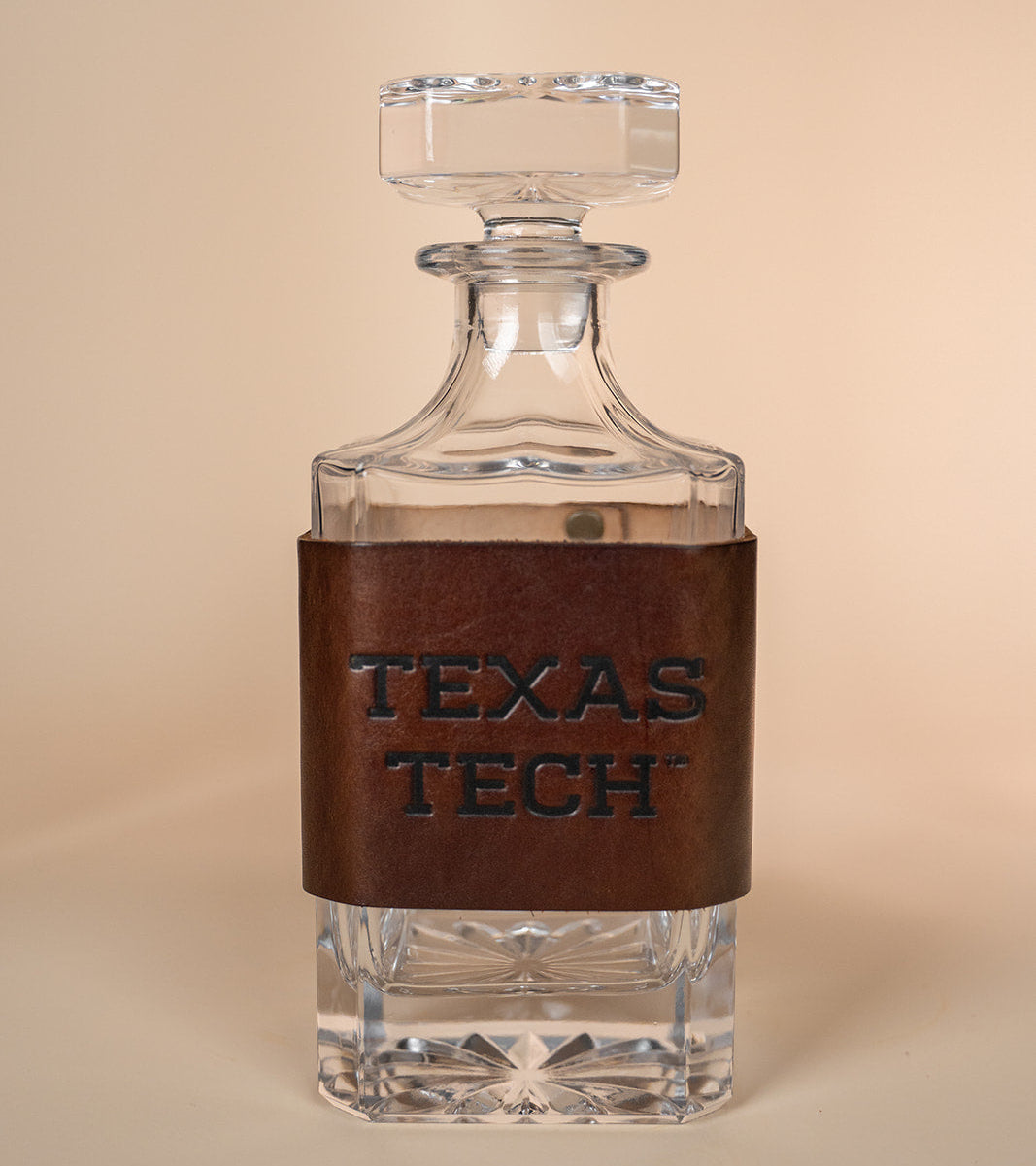 Clear glass decanter with brown leather wrap displaying 'Texas Tech' on a beige background