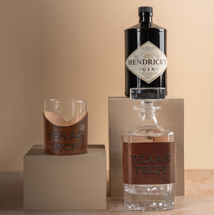Two lowball rocks glasses and a glass decanter with brown leather sleeves displaying &