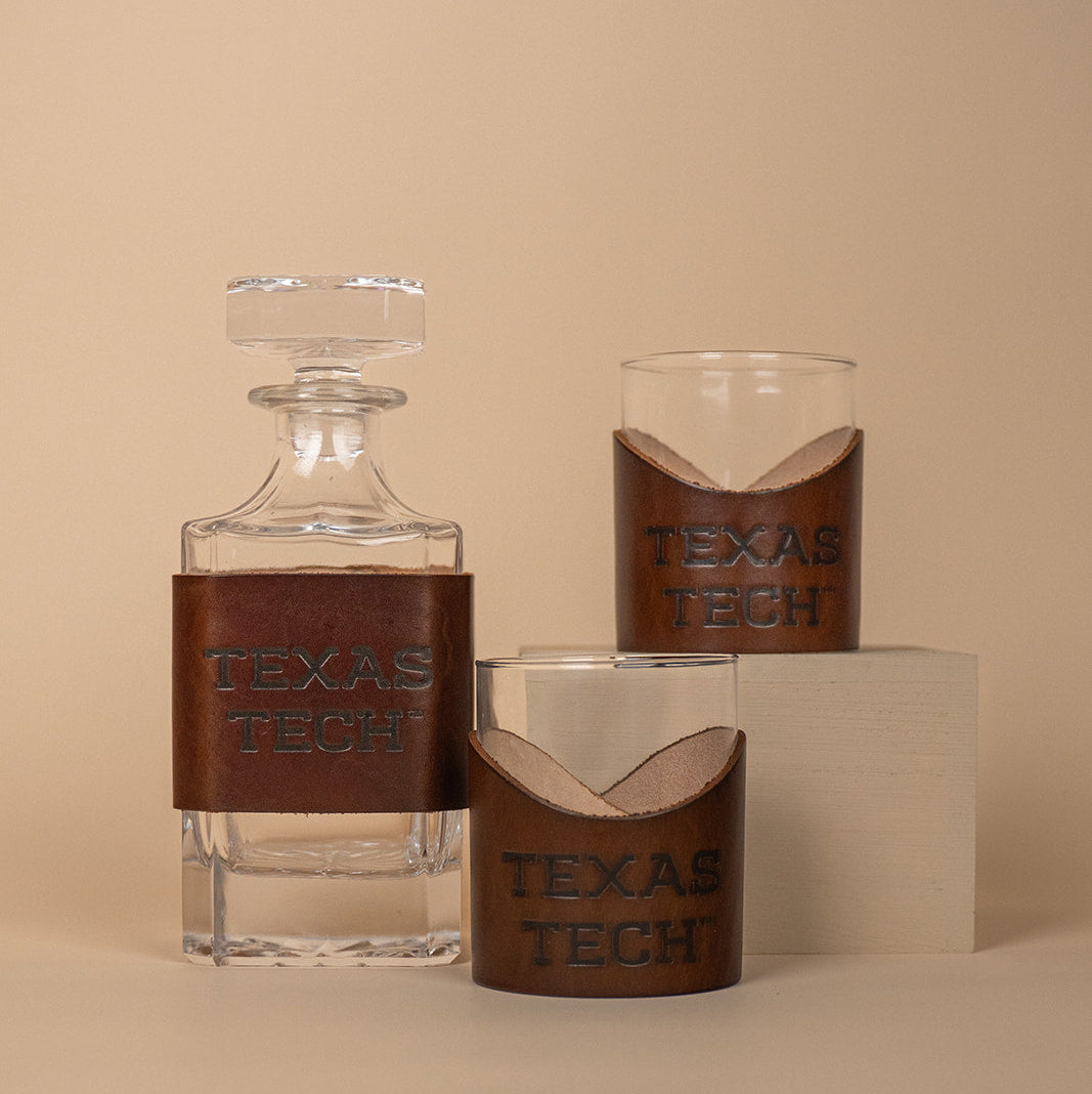 Two lowball rocks glasses and a glass decanter with brown leather sleeves displaying &