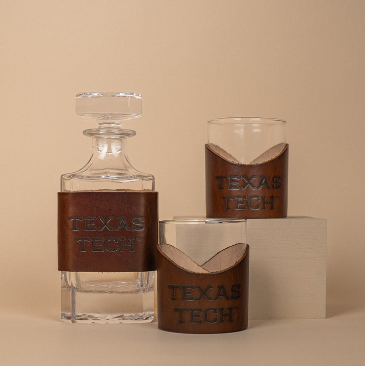 Two lowball rocks glasses and a glass decanter with brown leather sleeves displaying &