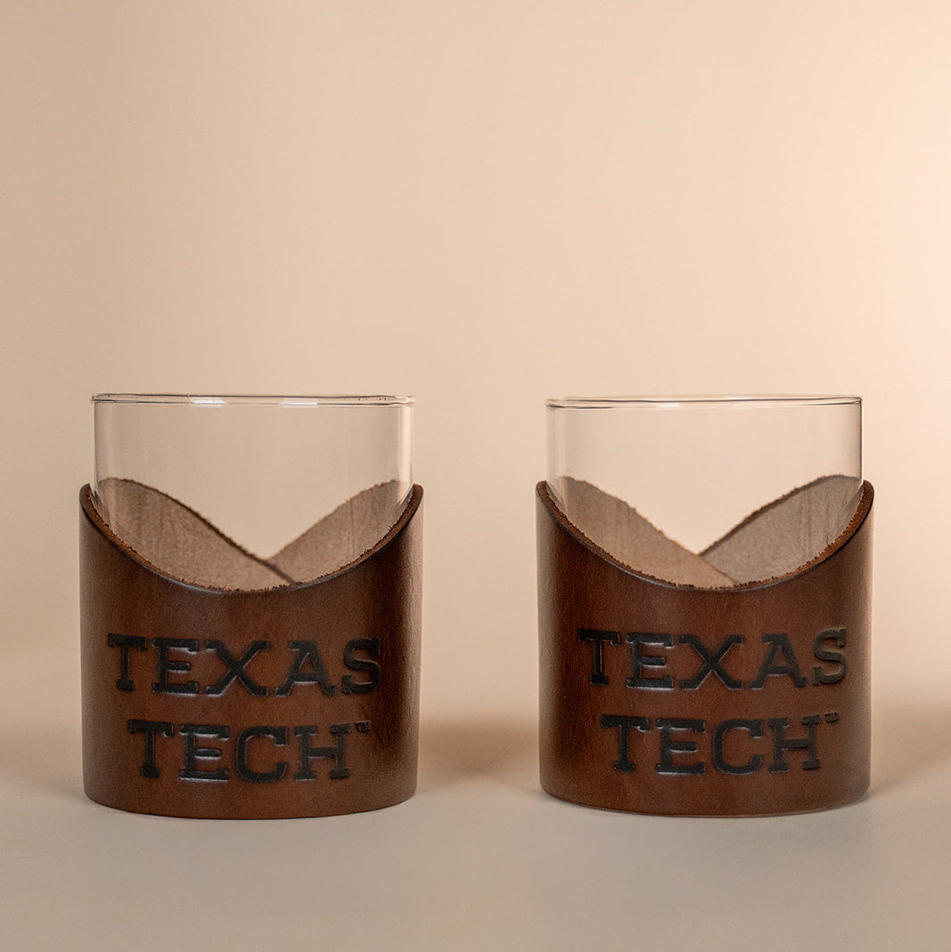 Two lowball rocks glasses with brown leather sleeves displaying 'TEXAS TECH' on a beige background.